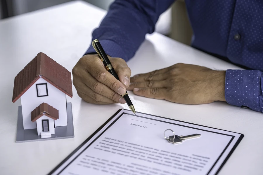 Man in a blue shirt signing a document beside a small house model and keys