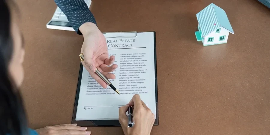 Overhead view of a real estate contract being signed beside a teal house model